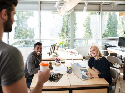 Image of happy young colleagues sitting in office coworking using laptop computers while their friend holding coffee. Looking aside.
